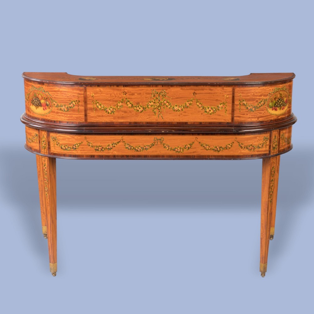 A hand-painted satinwood Carlton House desk featuring a curved superstructure of drawers, classical portrait medallions, floral swags, a leather writing surface, and square tapered legs on castors. View V