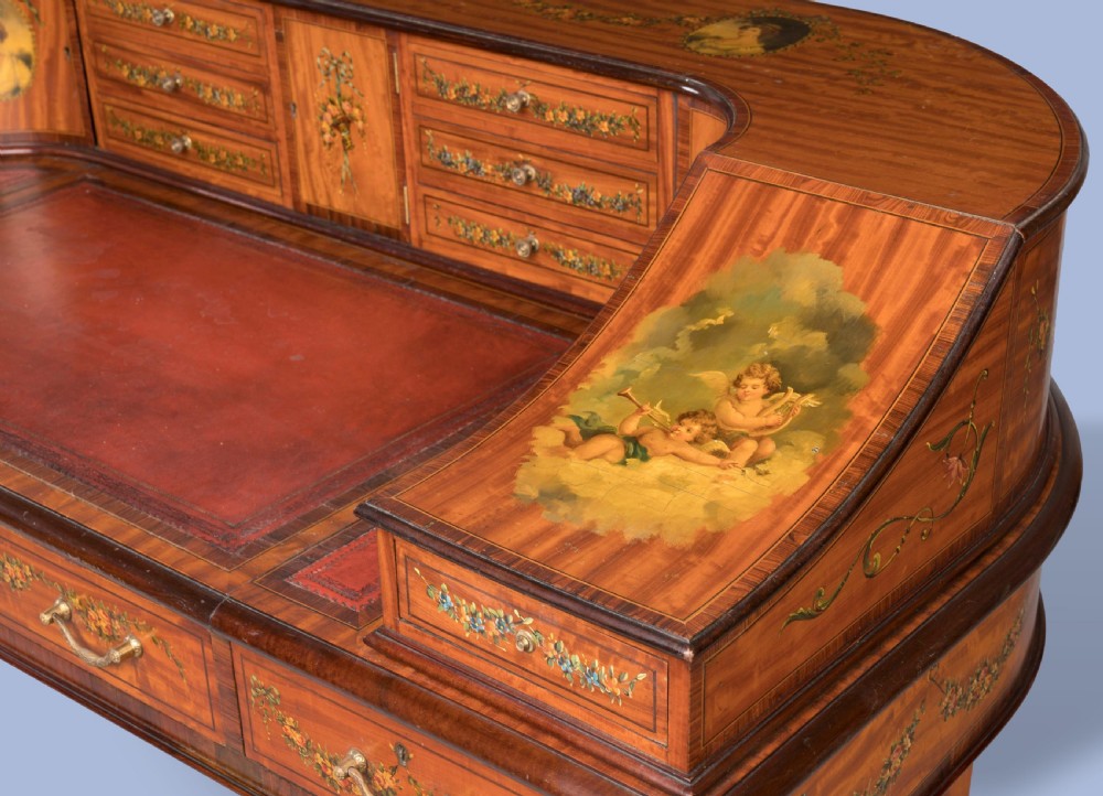 A hand-painted satinwood Carlton House desk featuring a curved superstructure of drawers, classical portrait medallions, floral swags, a leather writing surface, and square tapered legs on castors. View VII