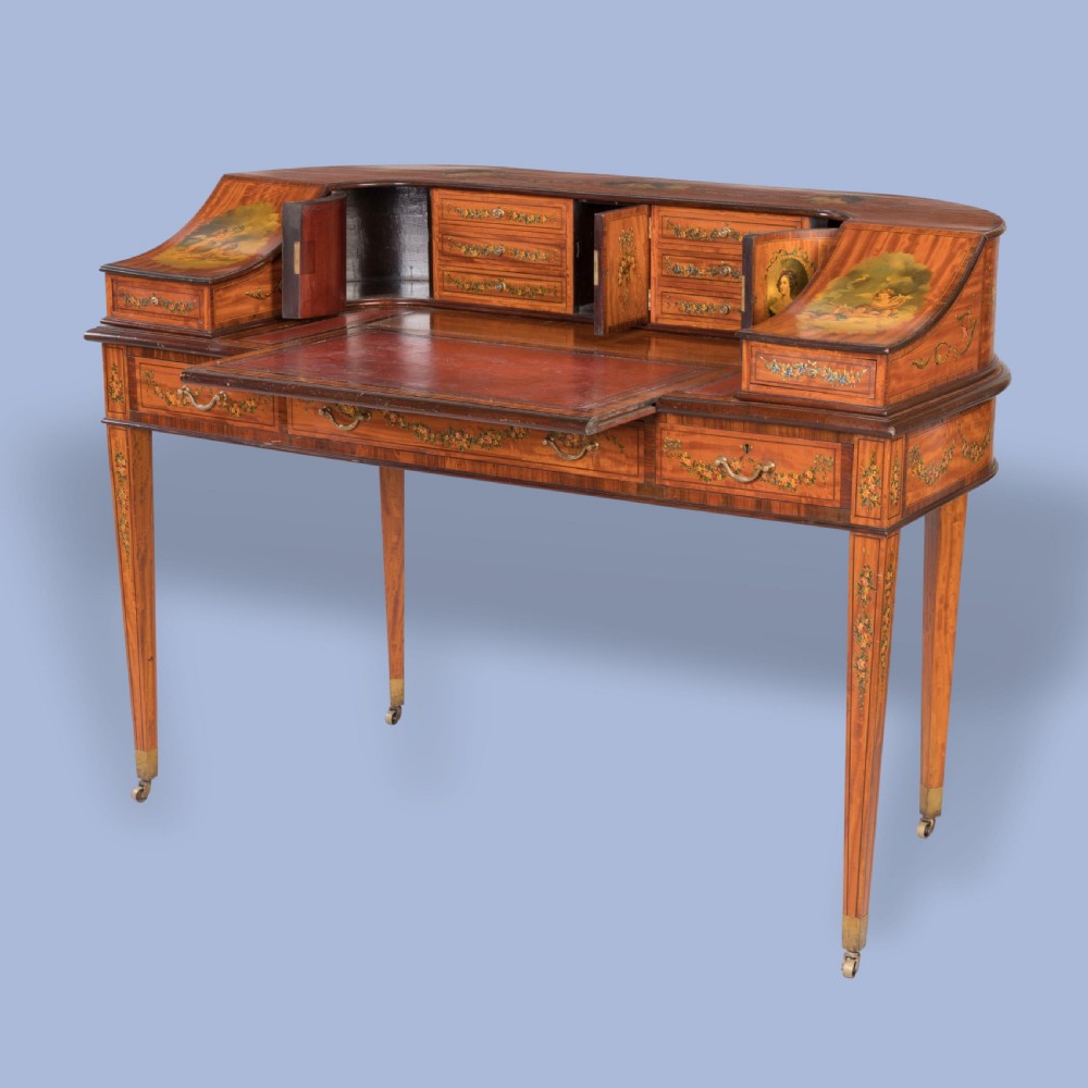 A hand-painted satinwood Carlton House desk featuring a curved superstructure of drawers, classical portrait medallions, floral swags, a leather writing surface, and square tapered legs on castors. View VIII