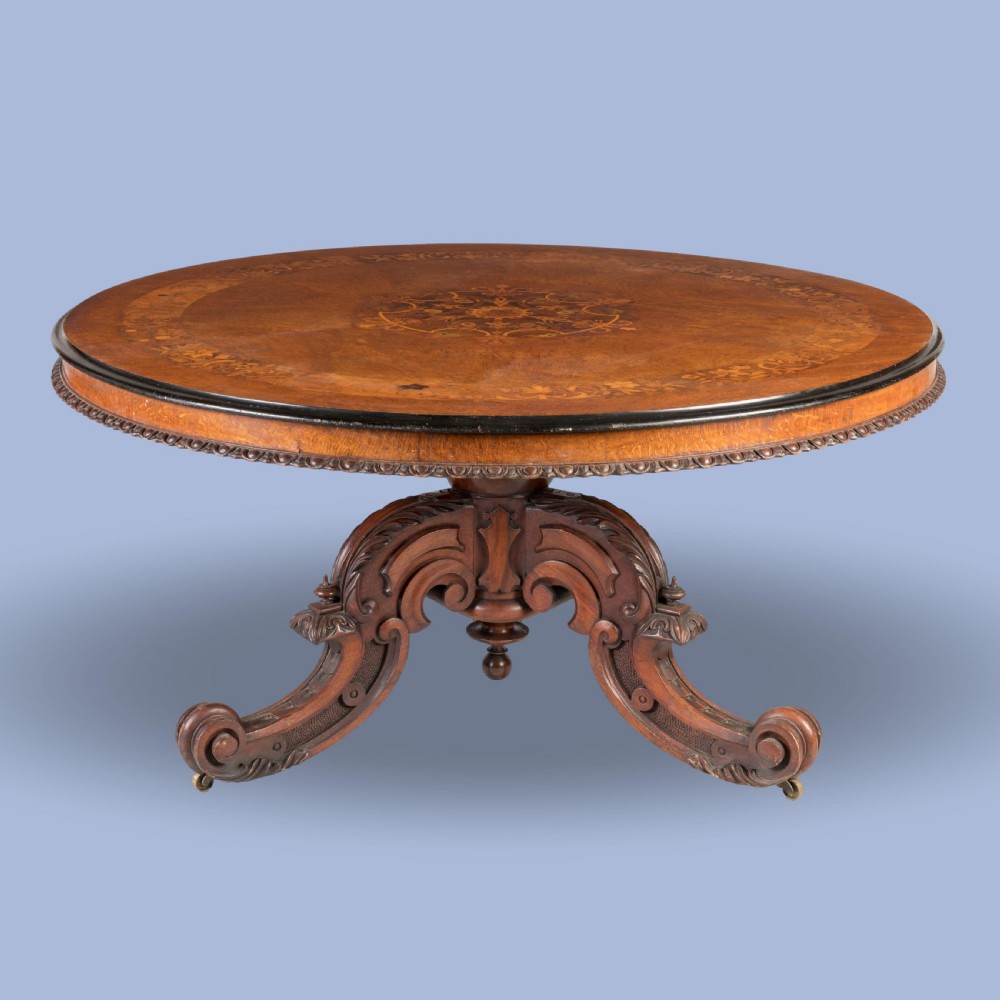 An early Victorian circular centre table in warm brown walnut, featuring decorative floral marquetry inlays on top and an intricately carved scroll-foot pedestal base.