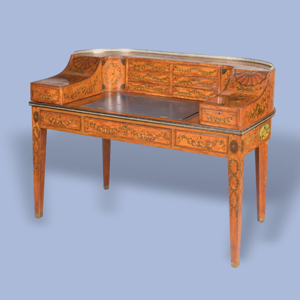 A satinwood Carlton House desk featuring hand-painted floral swags, a brass gallery rail, multiple superstructure drawers, and a central leather writing inset supported on square tapered legs.