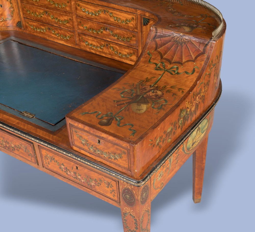 A satinwood Carlton House desk featuring hand-painted floral swags, a brass gallery rail, multiple superstructure drawers, and a central leather writing inset supported on square tapered legs. View VII