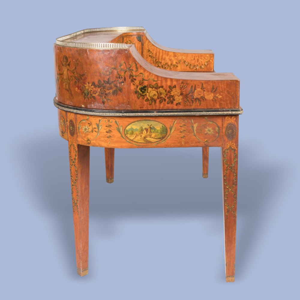 A satinwood Carlton House desk featuring hand-painted floral swags, a brass gallery rail, multiple superstructure drawers, and a central leather writing inset supported on square tapered legs. View IX