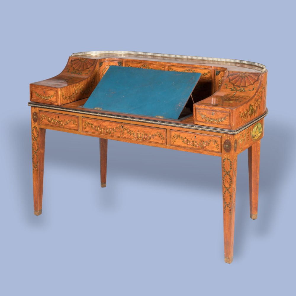 A satinwood Carlton House desk featuring hand-painted floral swags, a brass gallery rail, multiple superstructure drawers, and a central leather writing inset supported on square tapered legs. View X