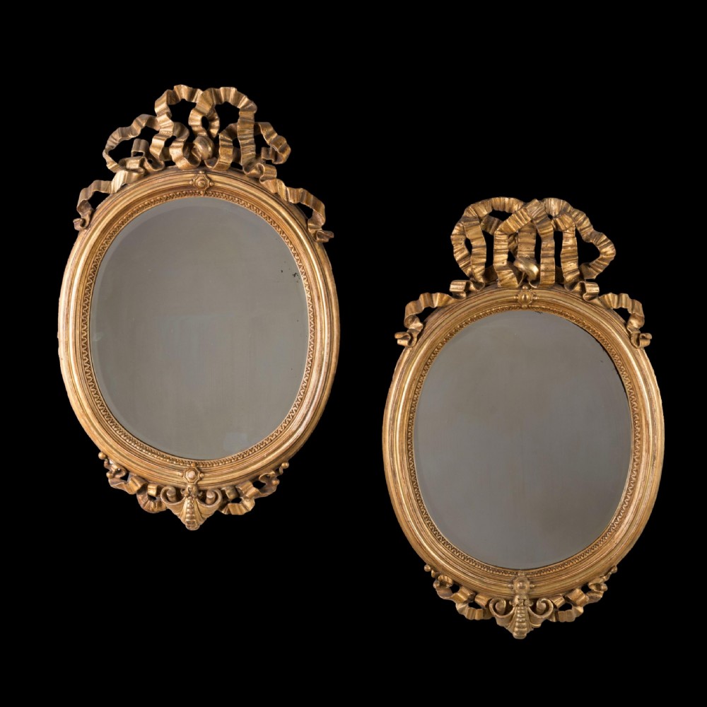 A matching pair of oval wall mirrors with gilded frames featuring intricate carved ribbon bow crestings.