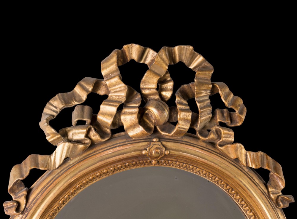A matching pair of oval wall mirrors with gilded frames featuring intricate carved ribbon bow crestings. View III