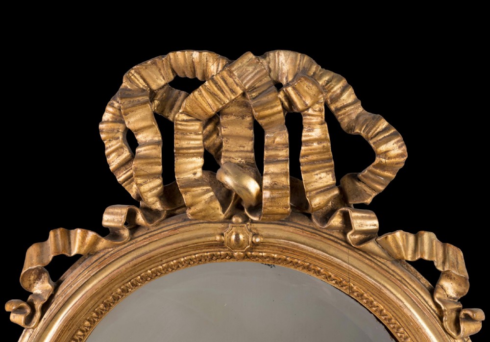A matching pair of oval wall mirrors with gilded frames featuring intricate carved ribbon bow crestings. View IV