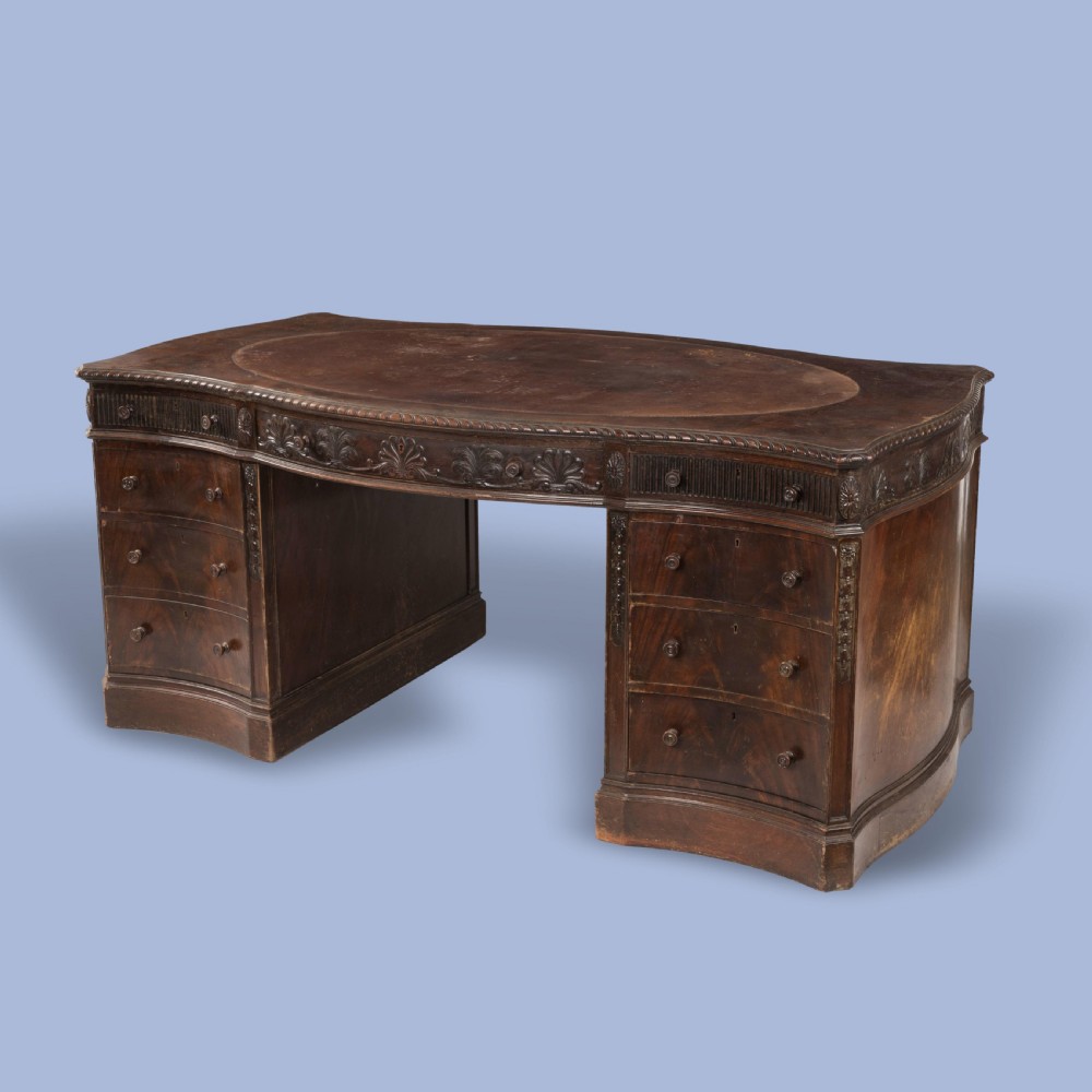 A dark mahogany serpentine pedestal desk featuring an oval leather-inset writing surface, carved foliate frieze, and twin drawer pedestals.