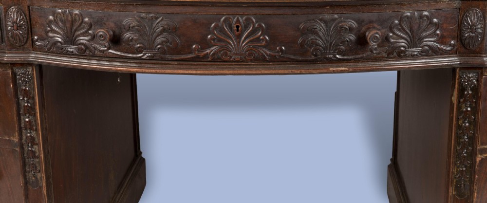 A dark mahogany serpentine pedestal desk featuring an oval leather-inset writing surface, carved foliate frieze, and twin drawer pedestals. View V