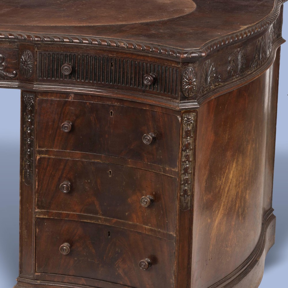 A dark mahogany serpentine pedestal desk featuring an oval leather-inset writing surface, carved foliate frieze, and twin drawer pedestals. View VI