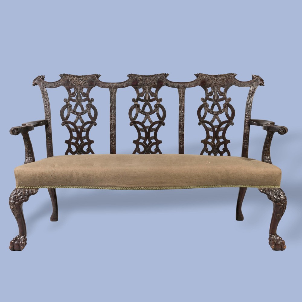 A Chippendale style dark mahogany triple chair-back settee featuring intricate pierced splats, carved cabriole legs, ball-and-claw feet, and a beige upholstered seat.