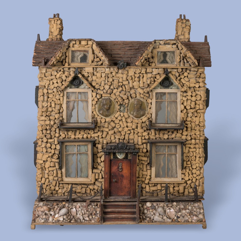 A miniature two-storey detached house constructed from wine corks and wood, featuring gables, glazed windows with lace curtains, classical medallion reliefs, and a base decorated with embedded seashells.
