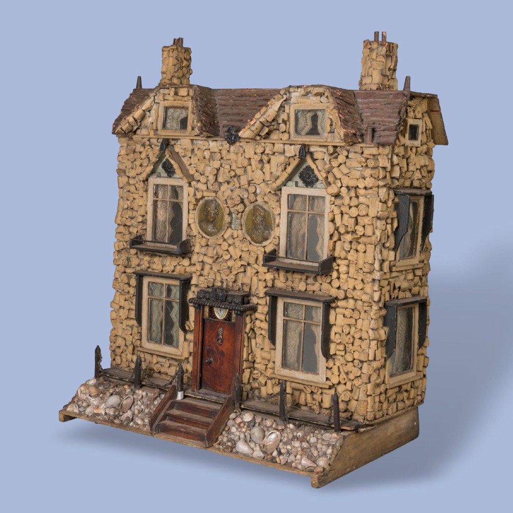 A miniature two-storey detached house constructed from wine corks and wood, featuring gables, glazed windows with lace curtains, classical medallion reliefs, and a base decorated with embedded seashells. View II
