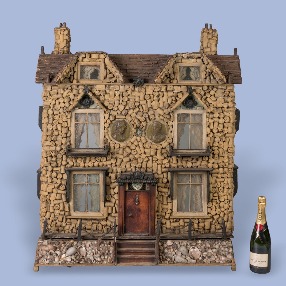 A miniature two-storey detached house constructed from wine corks and wood, featuring gables, glazed windows with lace curtains, classical medallion reliefs, and a base decorated with embedded seashells. View III