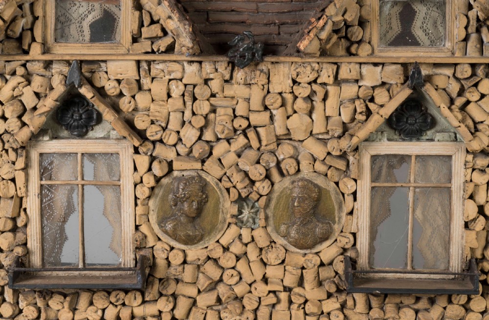A miniature two-storey detached house constructed from wine corks and wood, featuring gables, glazed windows with lace curtains, classical medallion reliefs, and a base decorated with embedded seashells. View VI