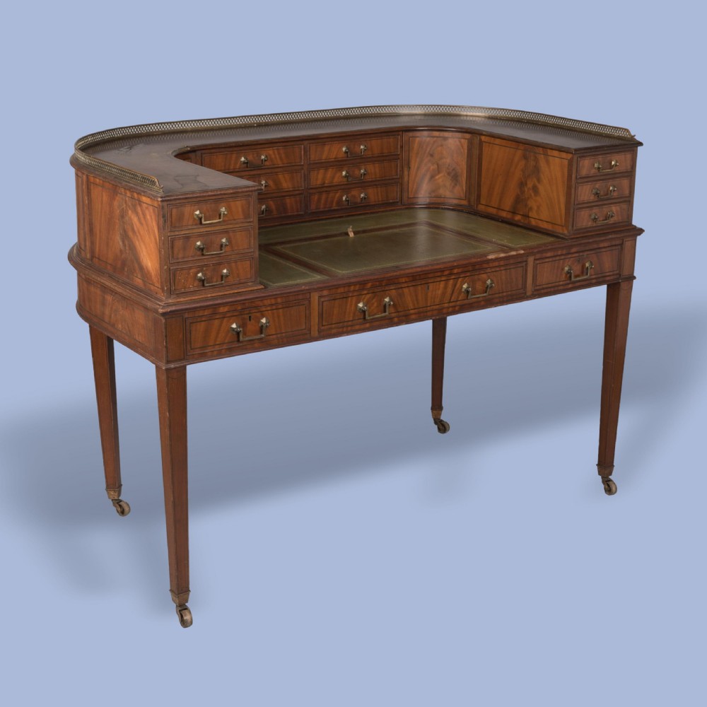 A mahogany Carlton House desk featuring a wrap-around drawer superstructure, brass gallery railing, green leather writing surface, and tapered legs on castors.