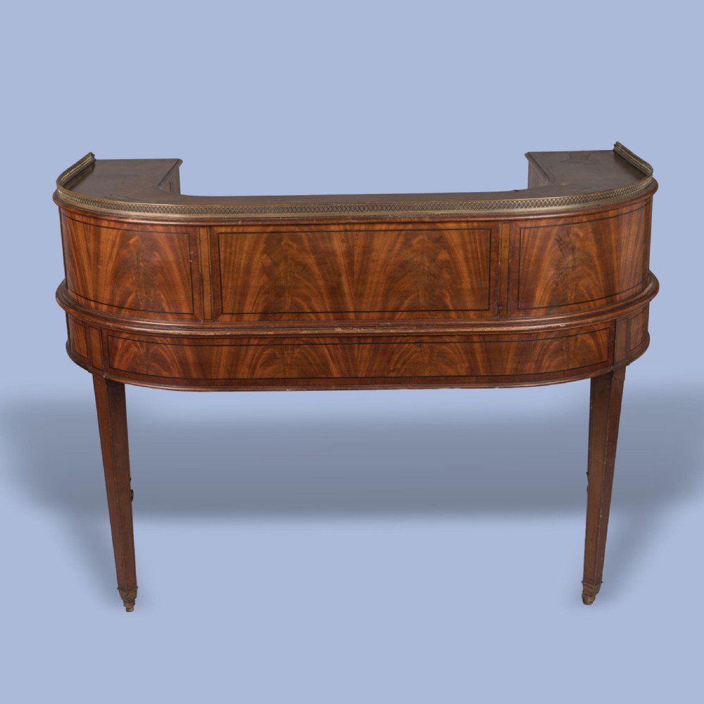 A mahogany Carlton House desk featuring a wrap-around drawer superstructure, brass gallery railing, green leather writing surface, and tapered legs on castors. View V