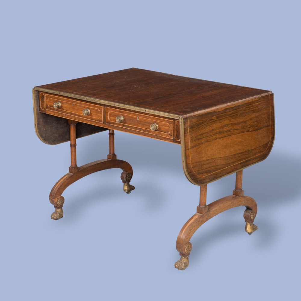 A Regency rosewood sofa table featuring drop leaves, two frieze drawers with brass knobs, brass edging, turned supports, and splayed legs ending in brass lion paw castors.