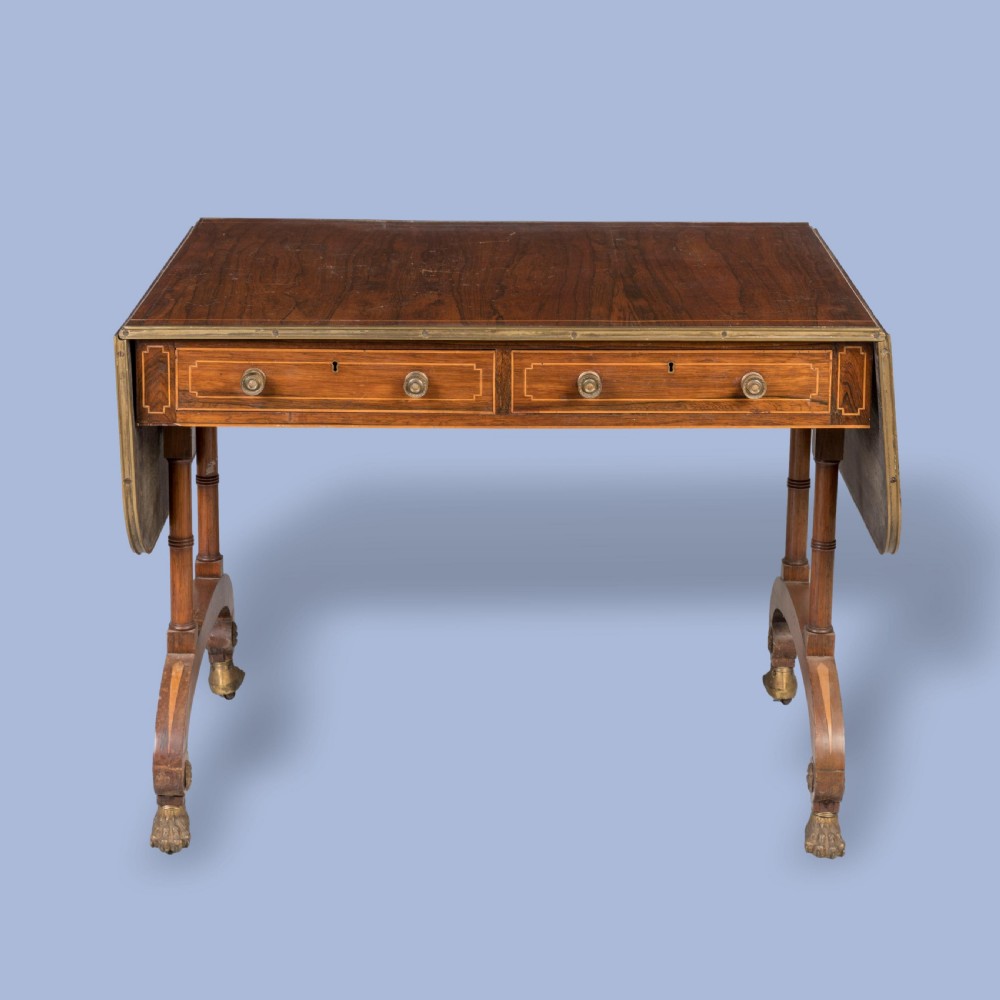 A Regency rosewood sofa table featuring drop leaves, two frieze drawers with brass knobs, brass edging, turned supports, and splayed legs ending in brass lion paw castors. View II