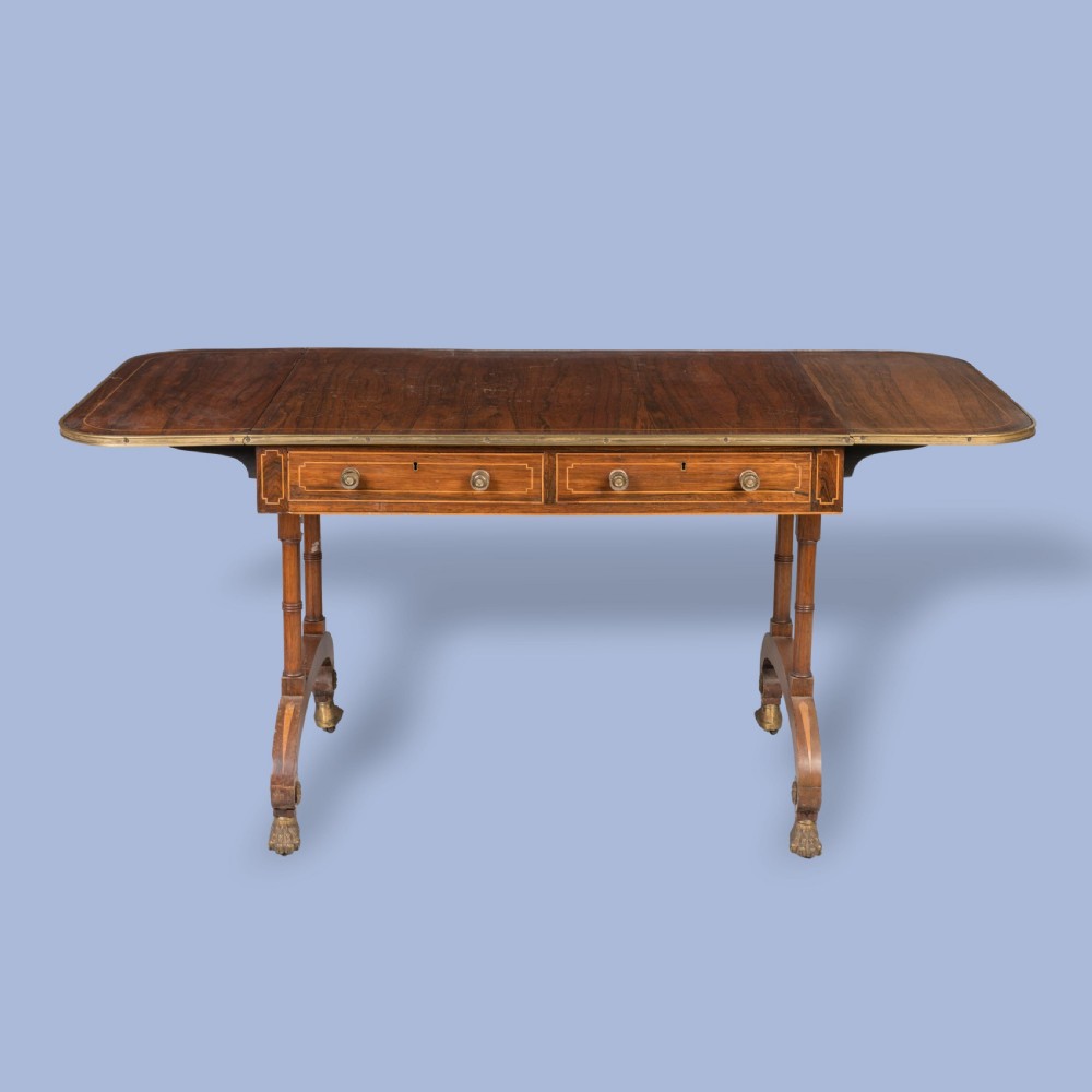 A Regency rosewood sofa table featuring drop leaves, two frieze drawers with brass knobs, brass edging, turned supports, and splayed legs ending in brass lion paw castors. View IV