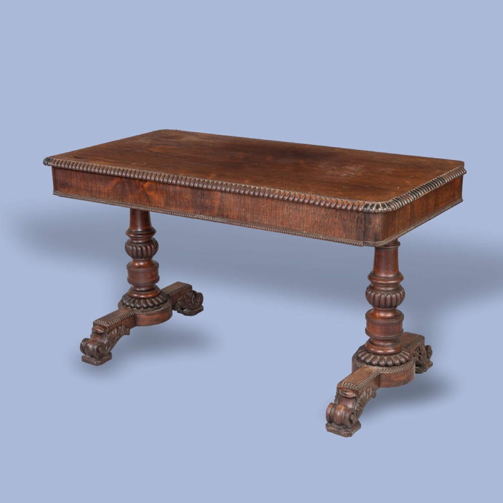 A Late Georgian rosewood writing table featuring a rectangular top with carved egg-and-dart moulding, resting on twin turned column pedestals and carved scroll feet.