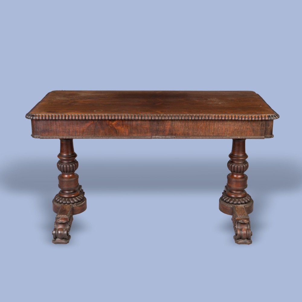A Late Georgian rosewood writing table featuring a rectangular top with carved egg-and-dart moulding, resting on twin turned column pedestals and carved scroll feet. View II