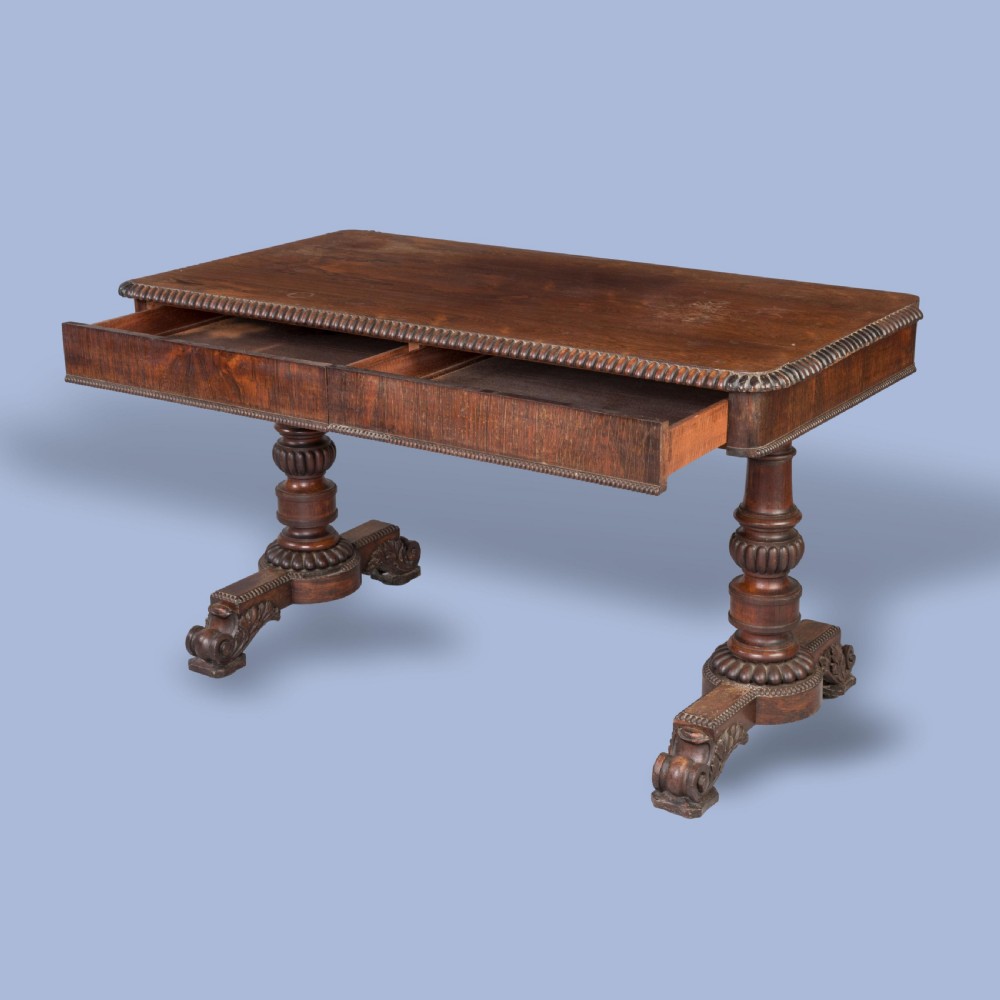 A Late Georgian rosewood writing table featuring a rectangular top with carved egg-and-dart moulding, resting on twin turned column pedestals and carved scroll feet. View III