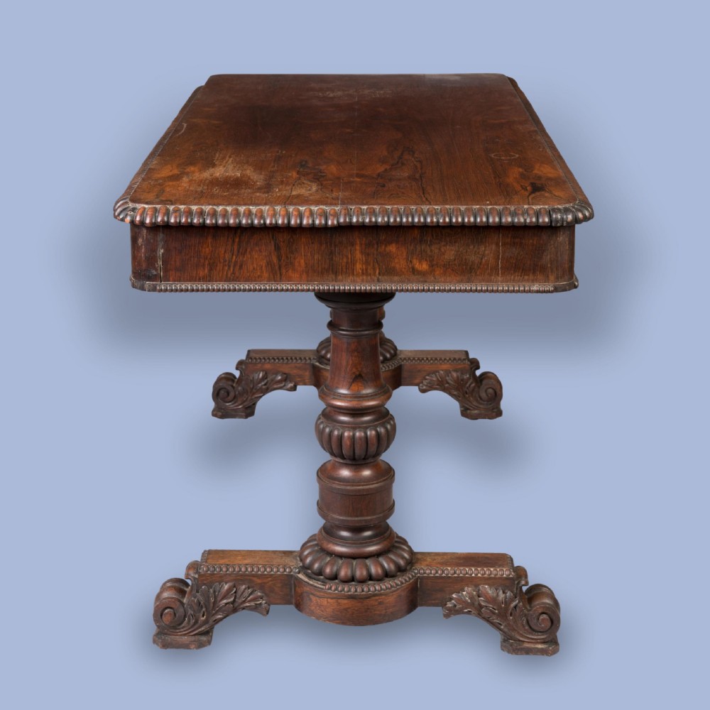 A Late Georgian rosewood writing table featuring a rectangular top with carved egg-and-dart moulding, resting on twin turned column pedestals and carved scroll feet. View V