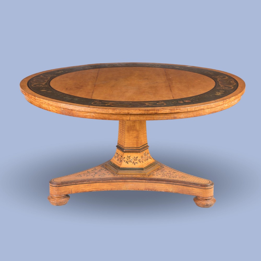 A Regency period circular centre table crafted from light sycamore with hand-painted floral borders resting on a central pedestal and triform base.