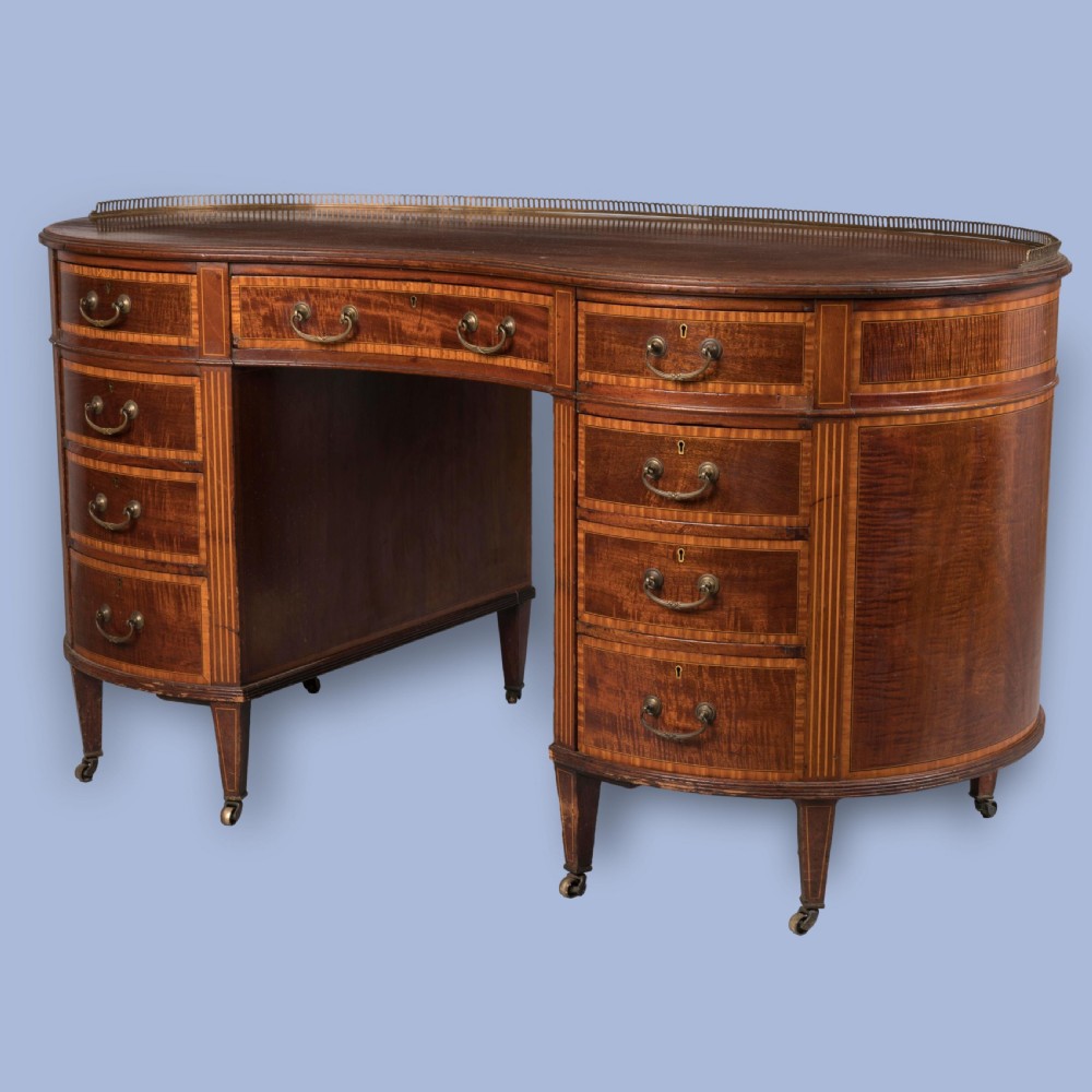 A late 19th-century mahogany kidney-shaped desk featuring satinwood inlay, brass drawer handles, a perforated brass top gallery, and tapered legs with brass castors.