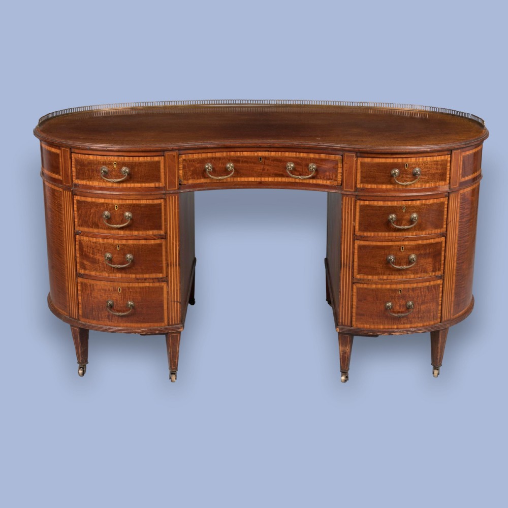 A late 19th-century mahogany kidney-shaped desk featuring satinwood inlay, brass drawer handles, a perforated brass top gallery, and tapered legs with brass castors. View II