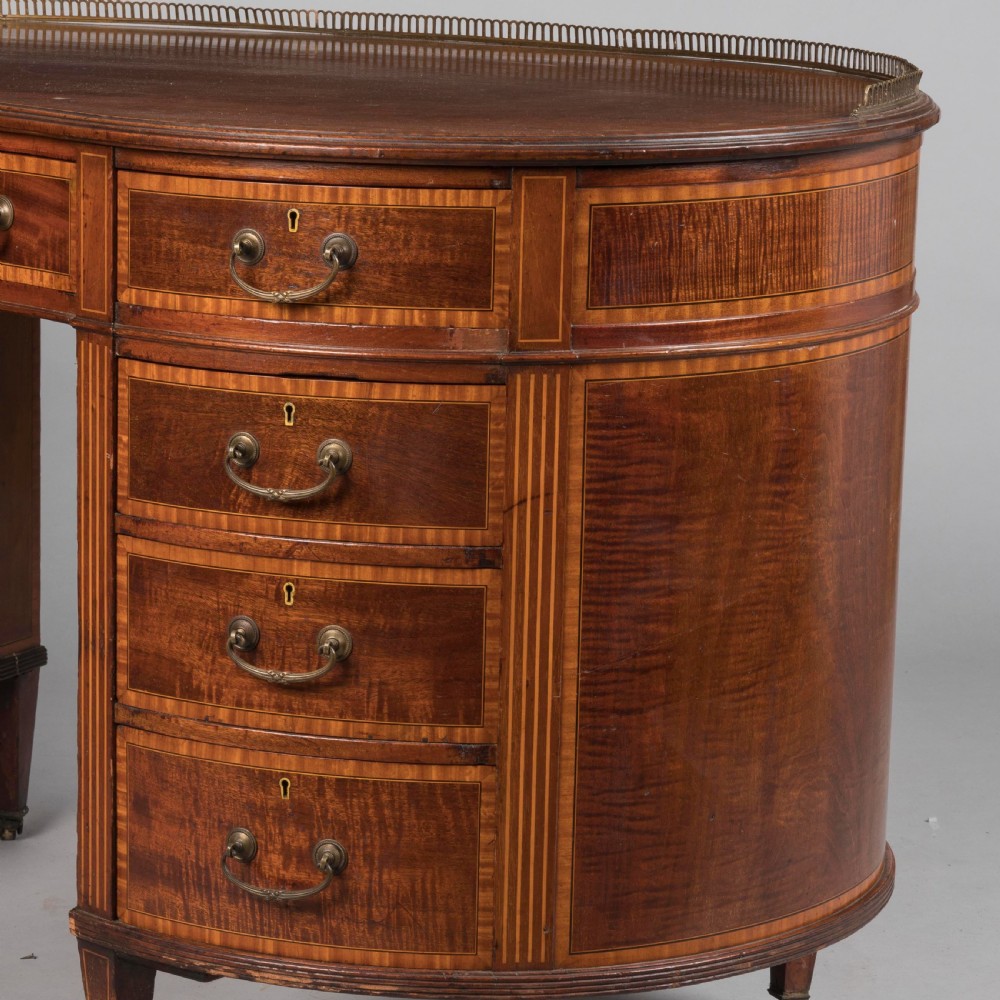A late 19th-century mahogany kidney-shaped desk featuring satinwood inlay, brass drawer handles, a perforated brass top gallery, and tapered legs with brass castors. View IV
