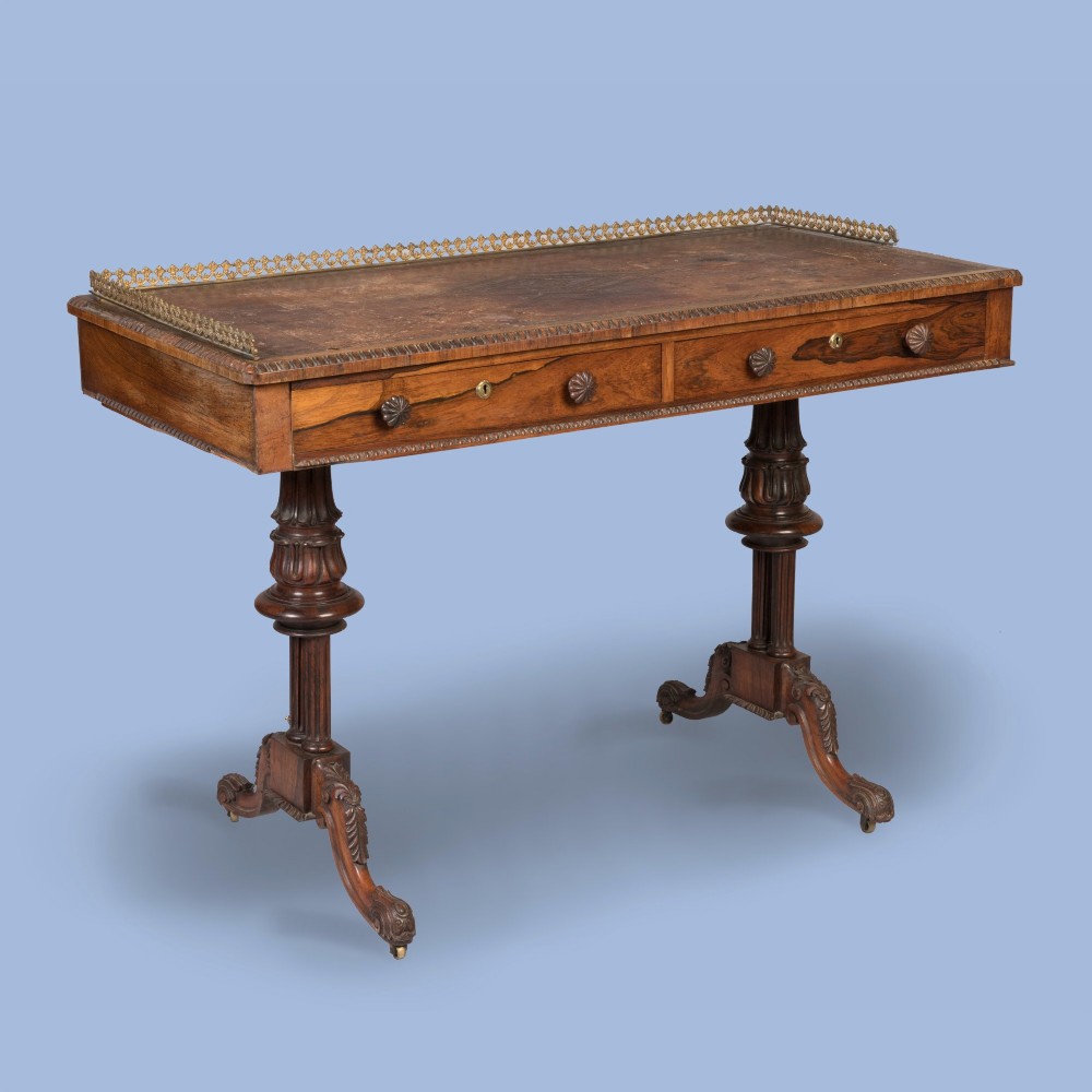 A Late Georgian rosewood writing table featuring a decorative brass gallery, an inset leather surface, two frieze drawers, and carved end supports resting on castors.
