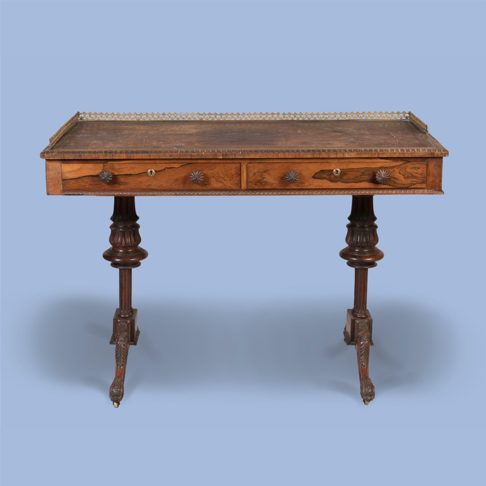 A Late Georgian rosewood writing table featuring a decorative brass gallery, an inset leather surface, two frieze drawers, and carved end supports resting on castors. View II