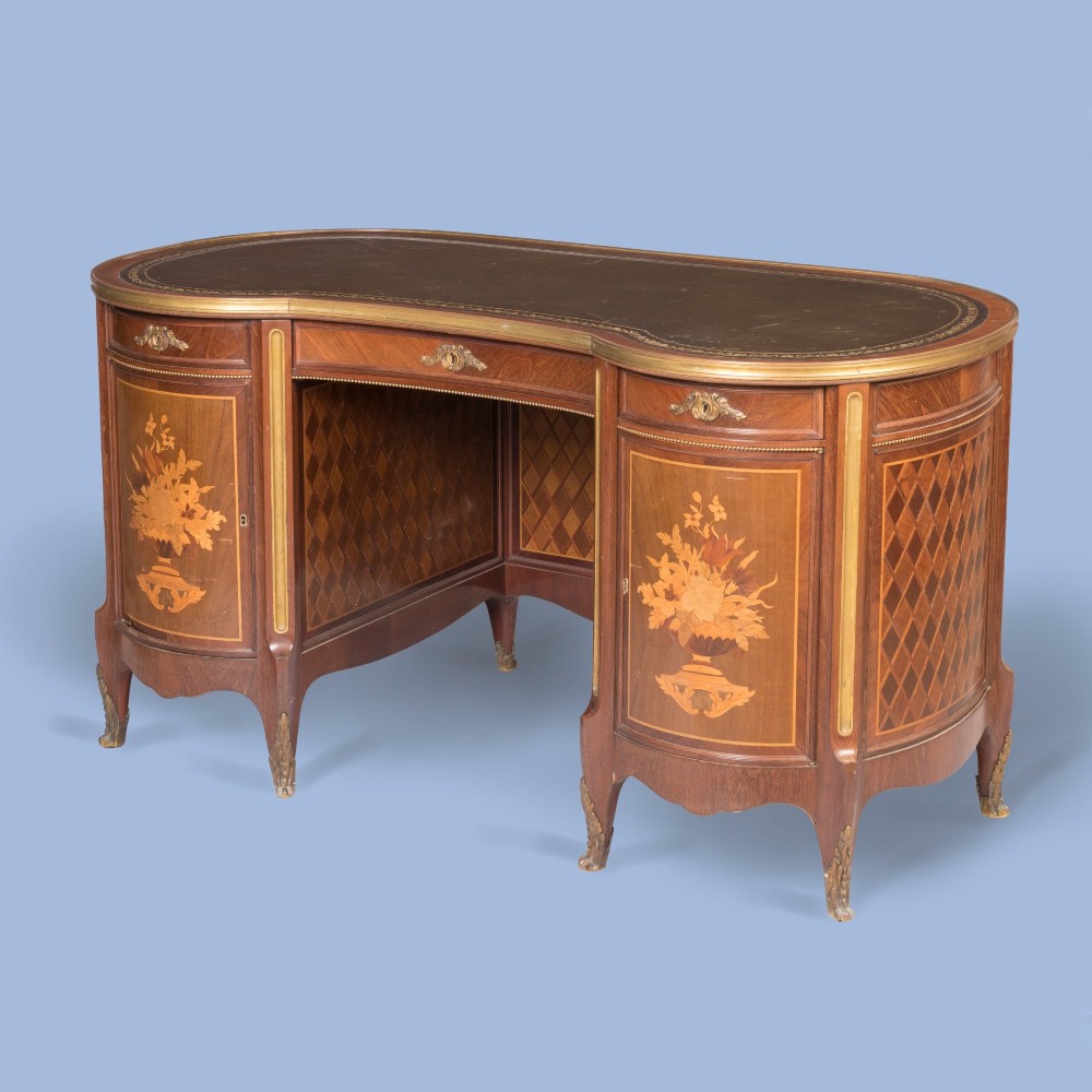 A kidney-shaped wooden writing desk featuring floral marquetry panels, gilt-bronze ormolu mounts, and a dark leather inset top with gold tooling.