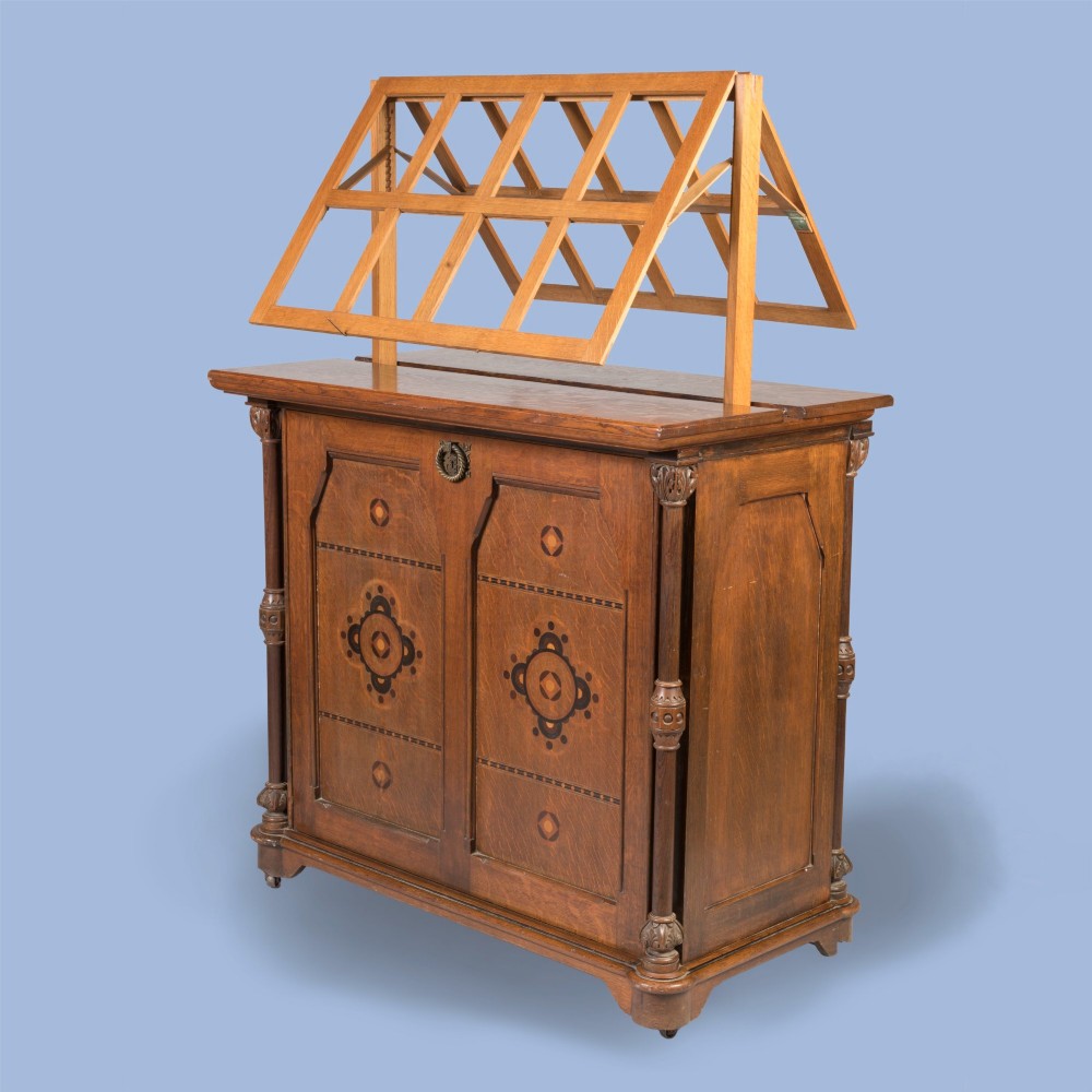 An antique Victorian Gothic Revival oak folio cabinet featuring geometric timber inlay on twin panel doors beneath an adjustable slatted display frame.