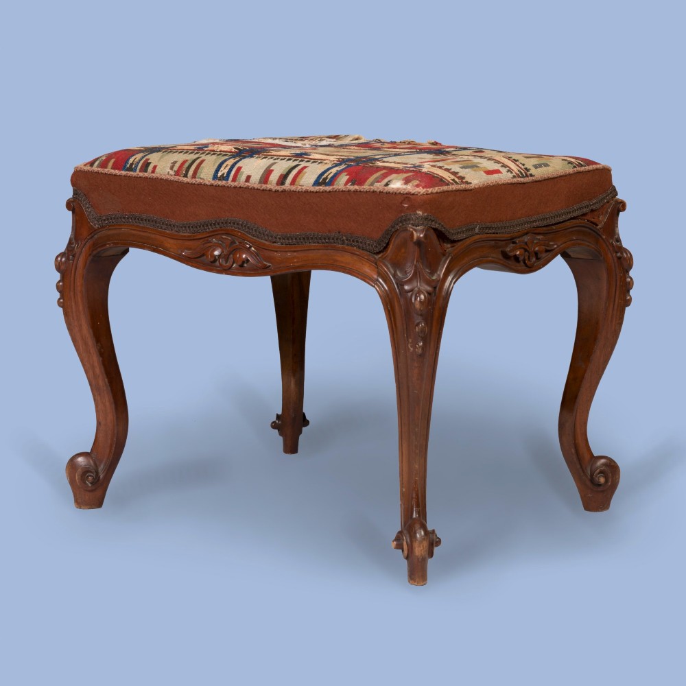 A carved walnut footstool featuring cabriole legs, scrolled feet, carved relief aprons, and a patterned tapestry seat.