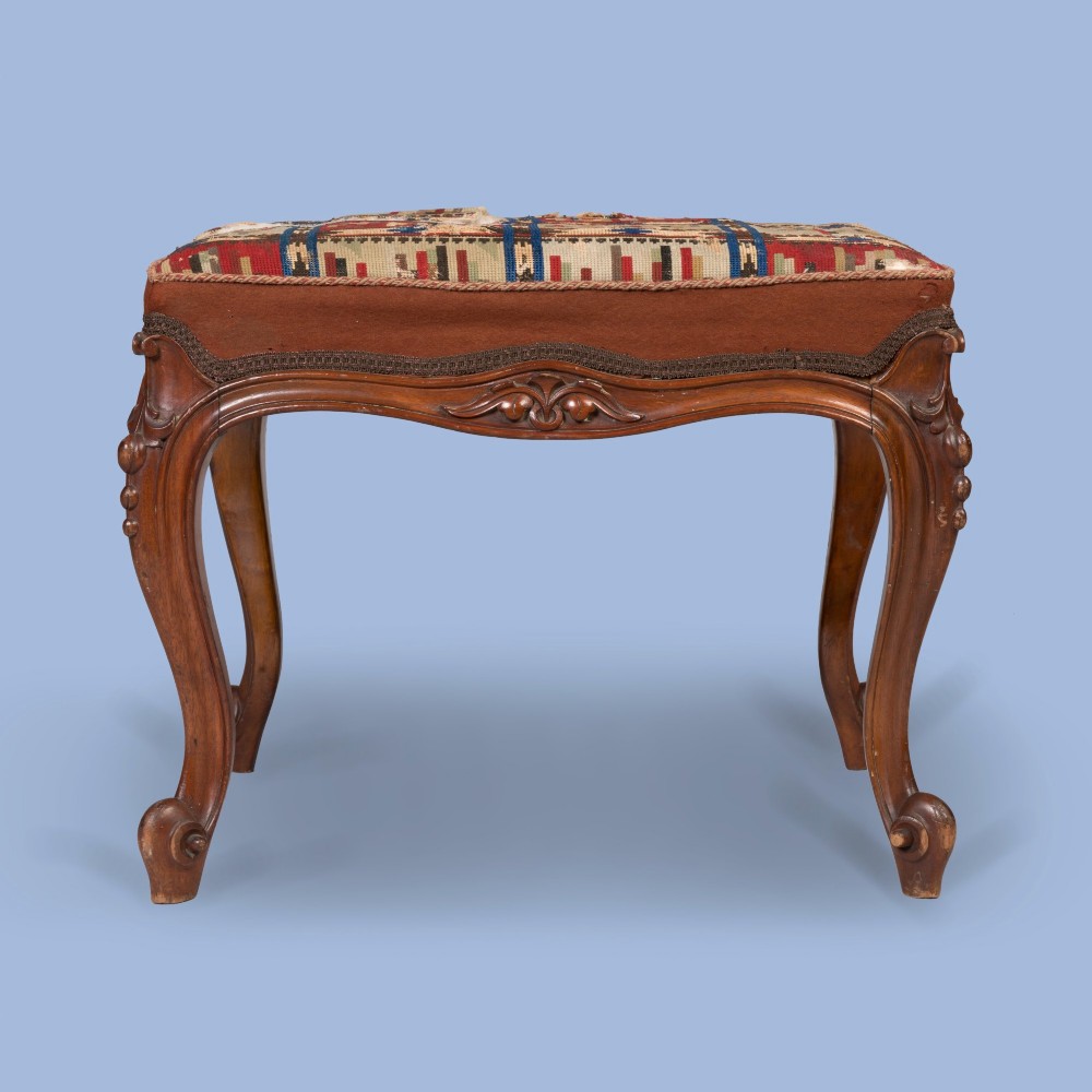 A carved walnut footstool featuring cabriole legs, scrolled feet, carved relief aprons, and a patterned tapestry seat. View II