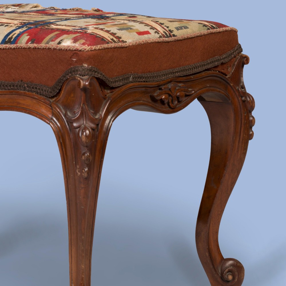 A carved walnut footstool featuring cabriole legs, scrolled feet, carved relief aprons, and a patterned tapestry seat. View III