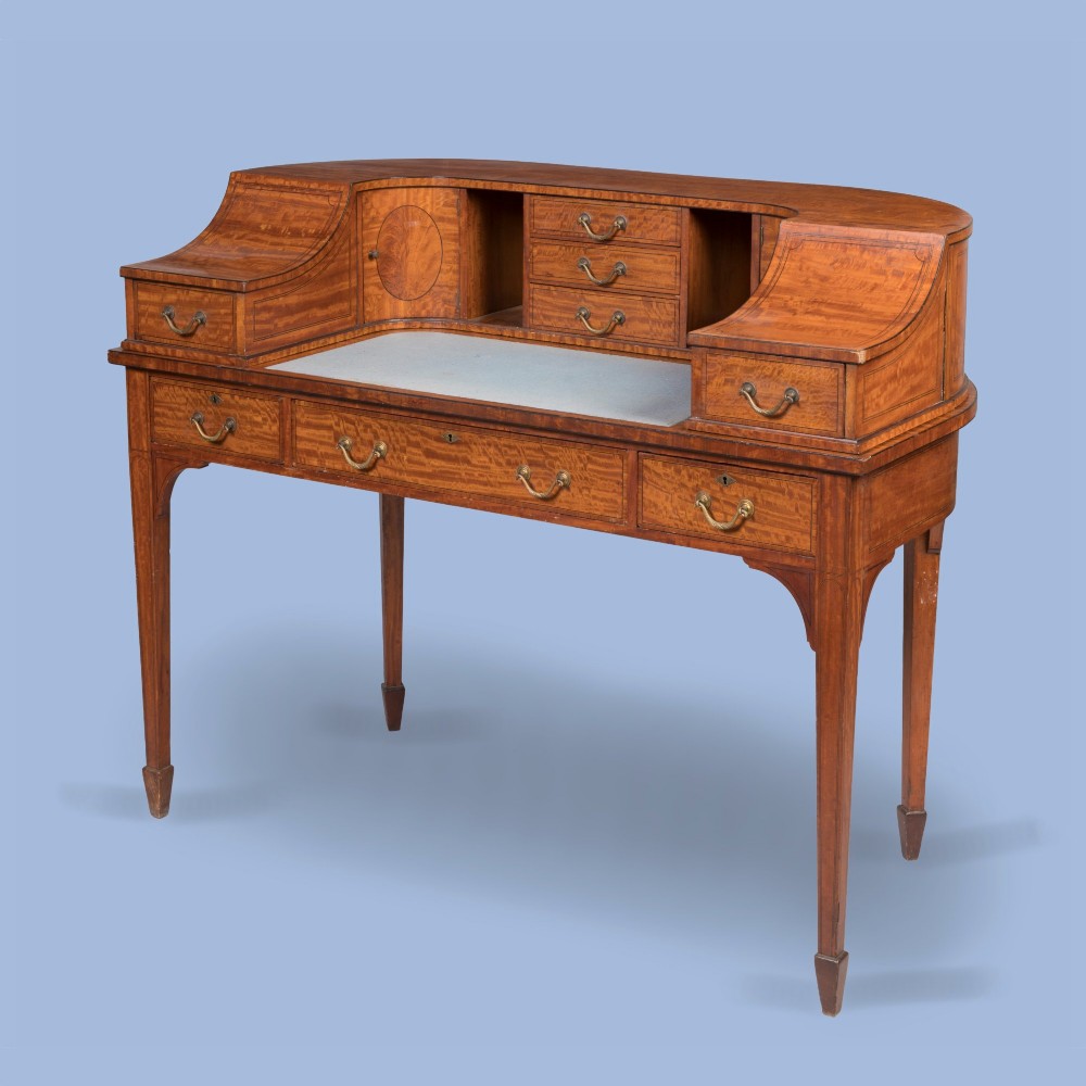 An Edwardian satinwood Carlton House desk featuring a curved rear superstructure with drawers, a leather writing surface, and square tapered legs.