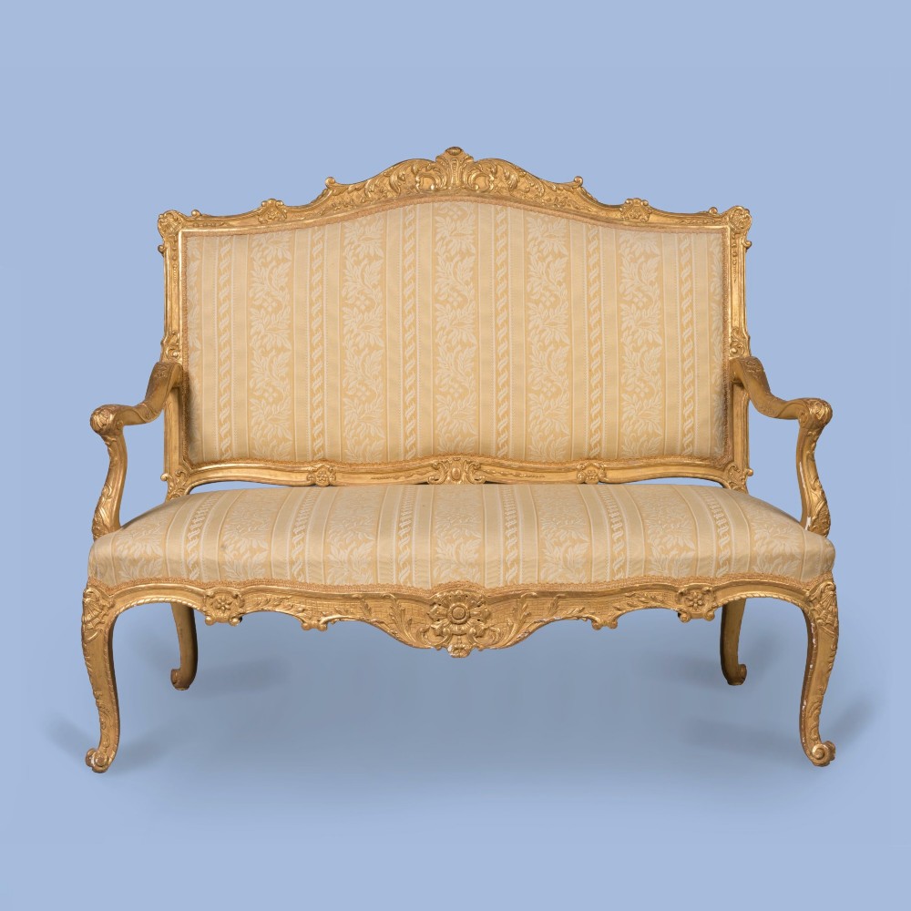 A French Louis XV style giltwood canape featuring carved floral cresting, cabriole legs, and light gold striped damask upholstery.
