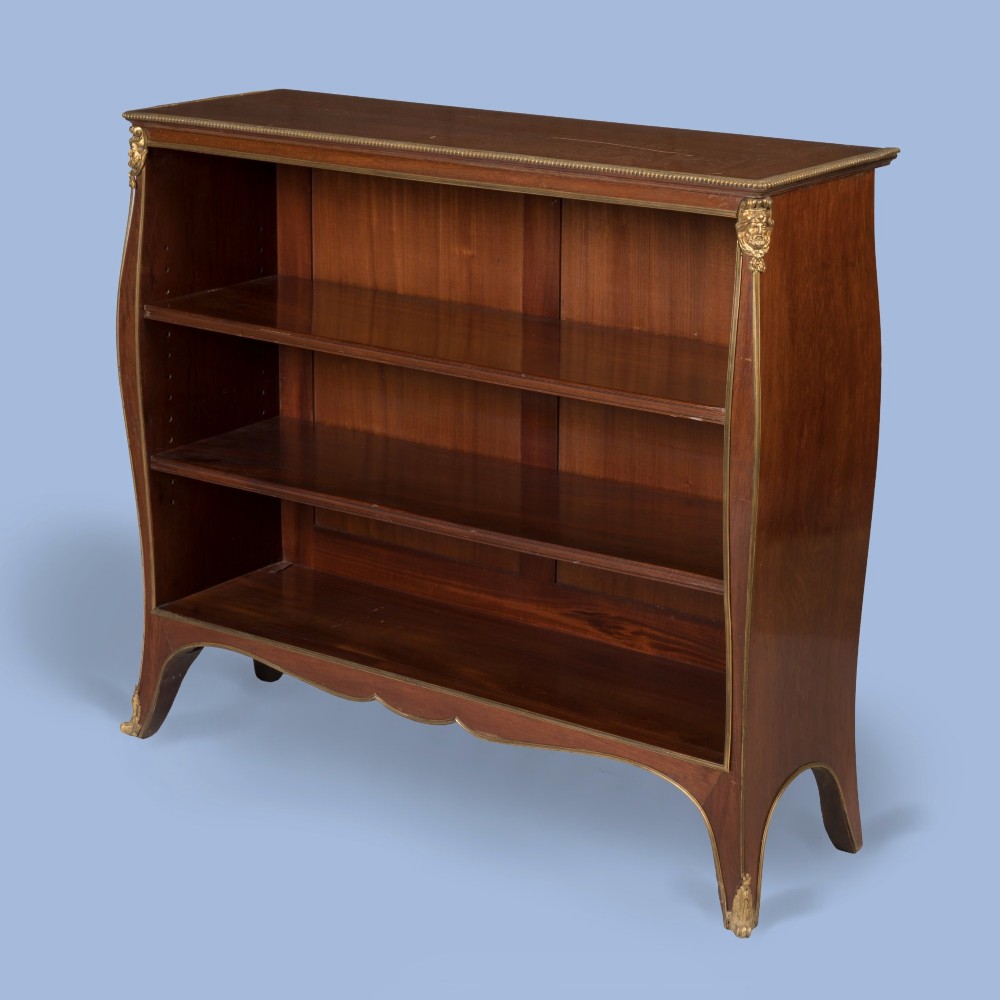 A low mahogany open bookcase featuring curved sides, adjustable shelves, a shaped apron, and gilt-bronze ormolu mounts on the corners and feet.