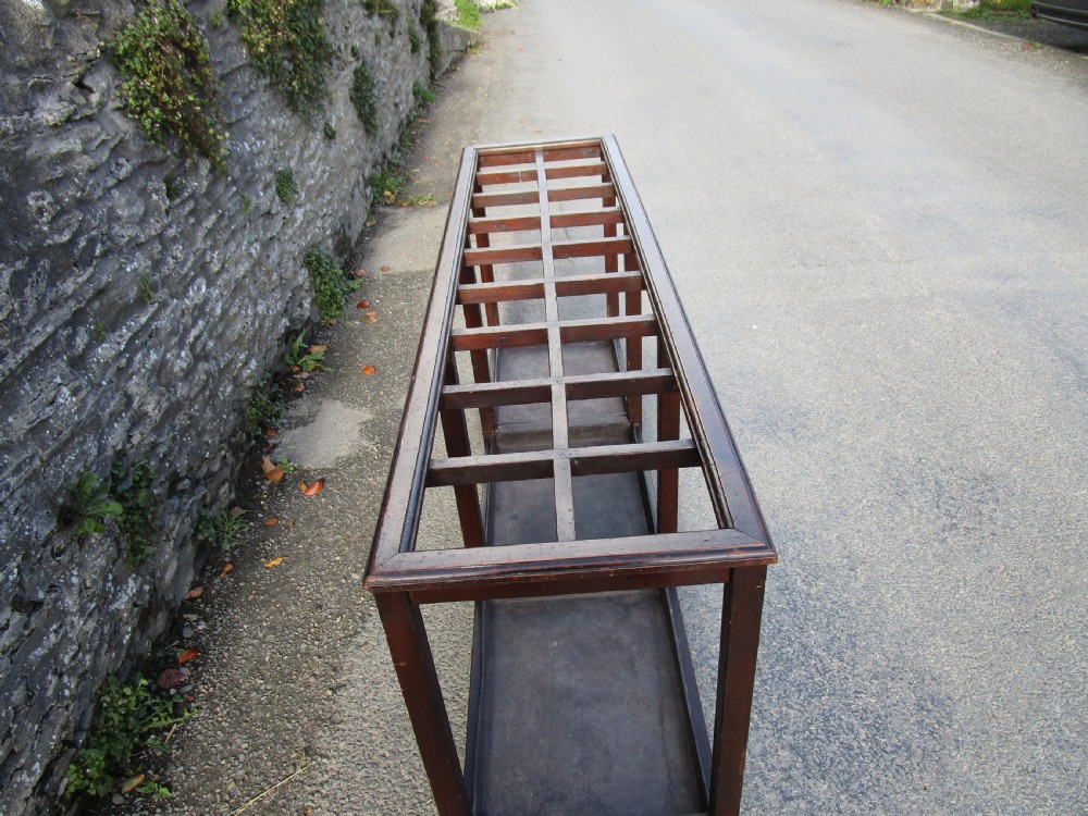 A long, dark wood stick stand featuring a grid of rectangular compartments at the top and sectioned metal drip trays at the base. View II
