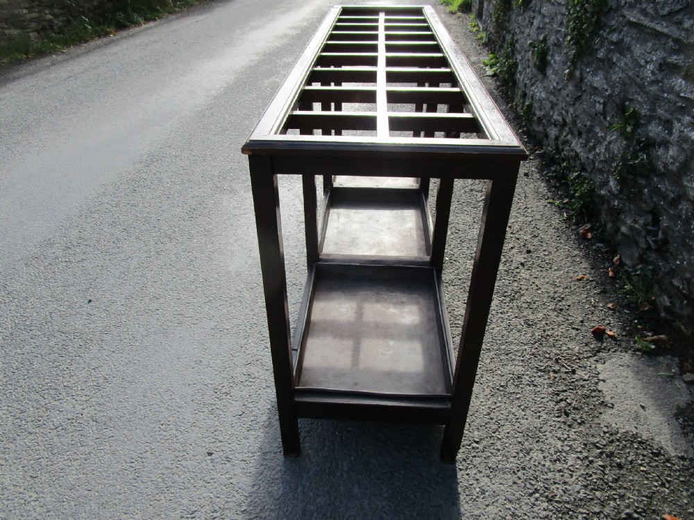 A long, dark wood stick stand featuring a grid of rectangular compartments at the top and sectioned metal drip trays at the base. View III