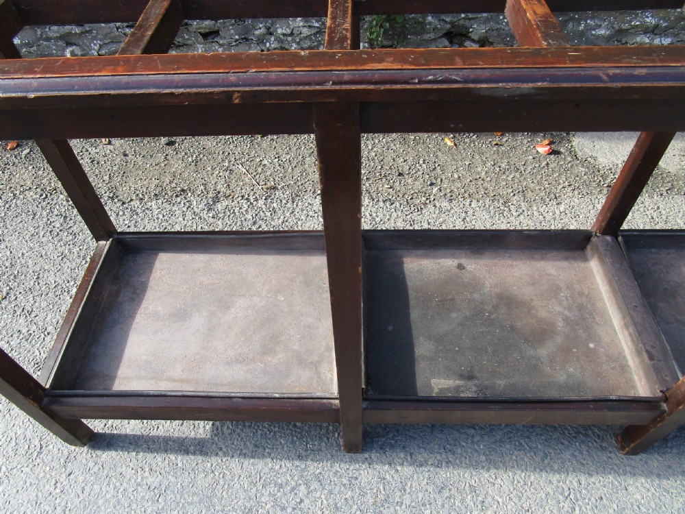 A long, dark wood stick stand featuring a grid of rectangular compartments at the top and sectioned metal drip trays at the base. View V