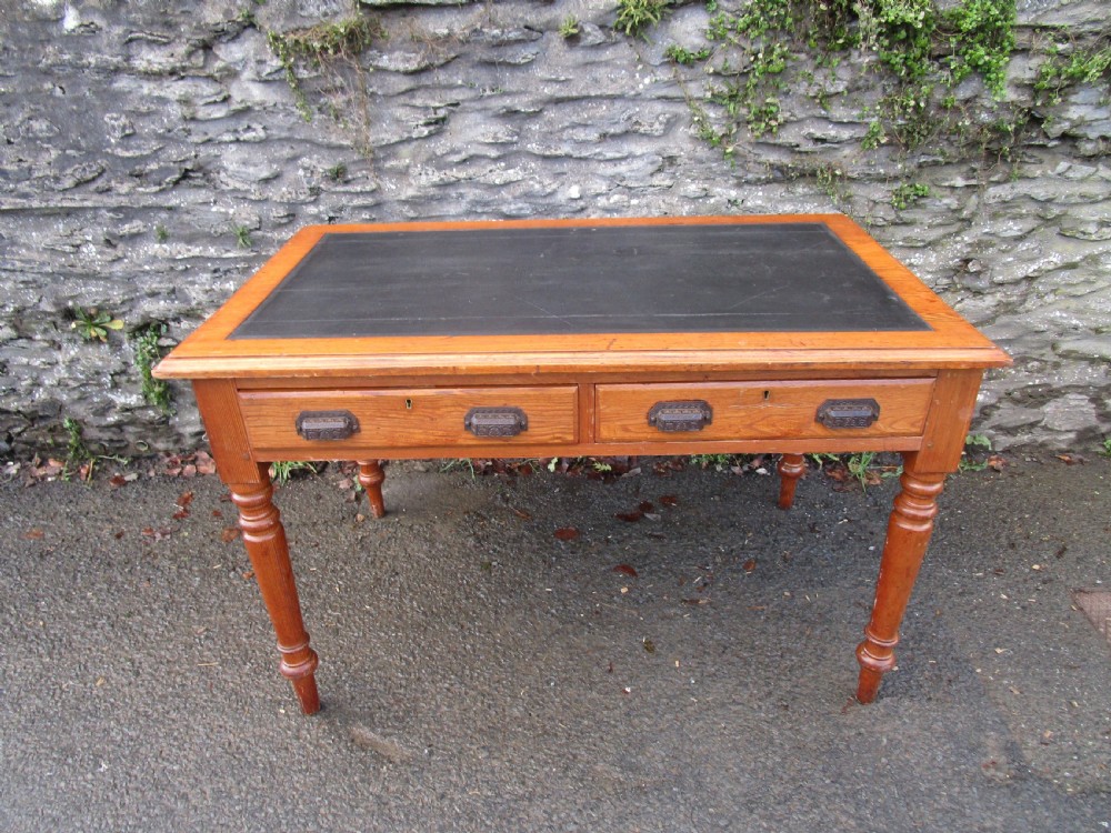 A warm brown pine writing table featuring a dark leather insert writing surface, two frieze drawers with decorative metal handles, and four turned legs.