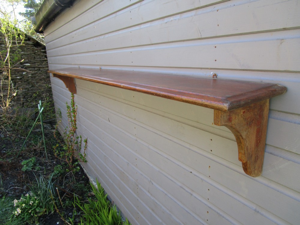 A long Victorian warm brown wooden hanging shelf with two curved supporting side brackets mounted against a timber wall. View II