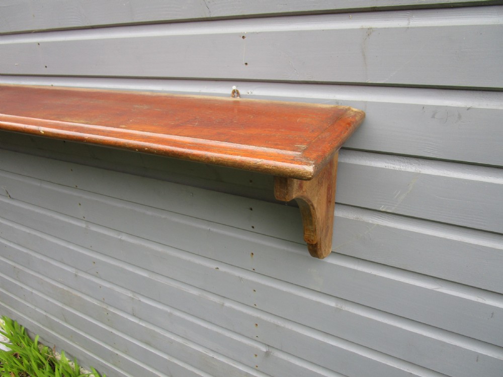 A long Victorian warm brown wooden hanging shelf with two curved supporting side brackets mounted against a timber wall. View III