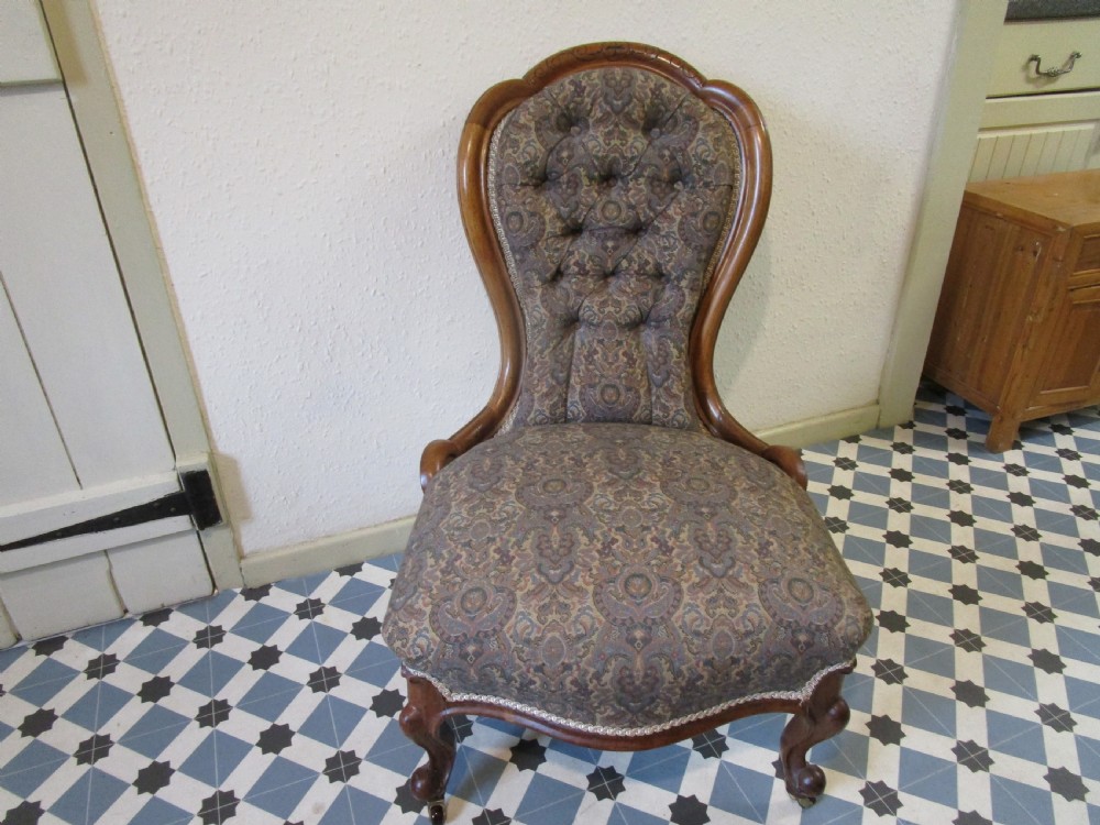 An antique Victorian low-seated nursing chair featuring a carved mahogany spoon back frame, buttoned upholstery in a patterned fabric, and cabriole front legs with brass castors.