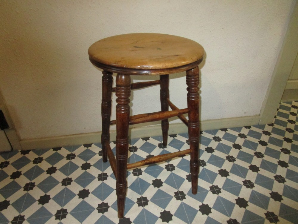 A rustic antique wooden stool featuring a circular ash top supported by four dark turned legs with horizontal stretchers.